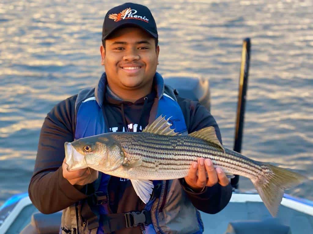 A photo of an angler holding a Striped Bass with both hands while standing on a charter fishing boat 