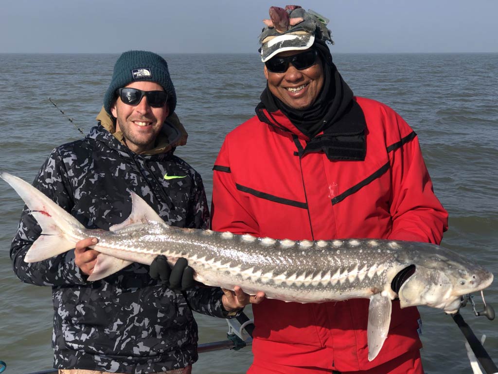 A photo of two anglers holding Sturgeon together while standing on a charter fishing boat on a river