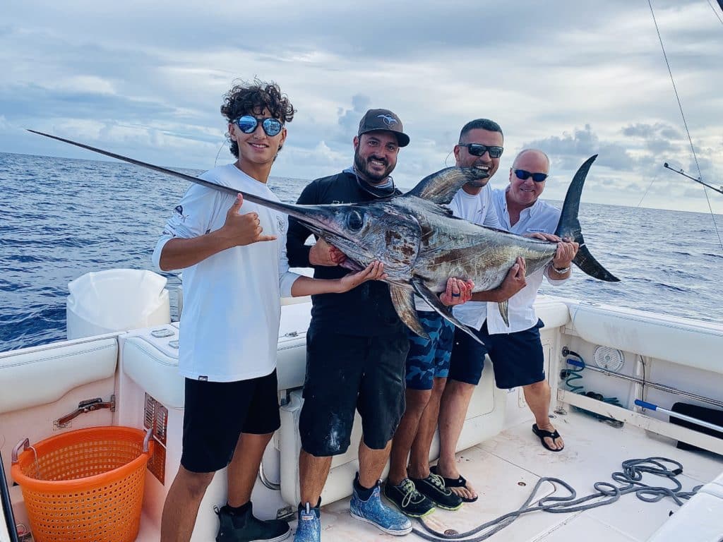 A group of four male anglers, all smiling and holding a freshly caught Swordfish while standing on a fishing boat in Miami Beach, FL