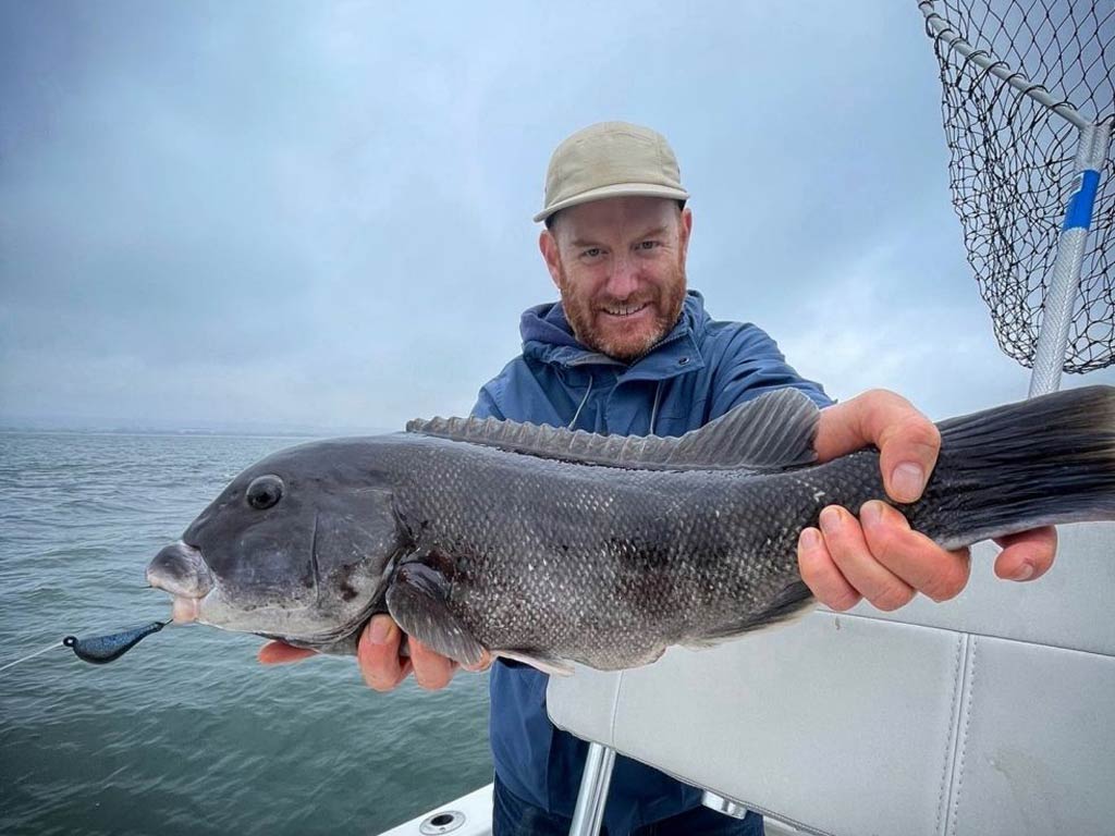 A photo of an angler standing on a boat and holding Tautog with both hands during a cloudy and cold day