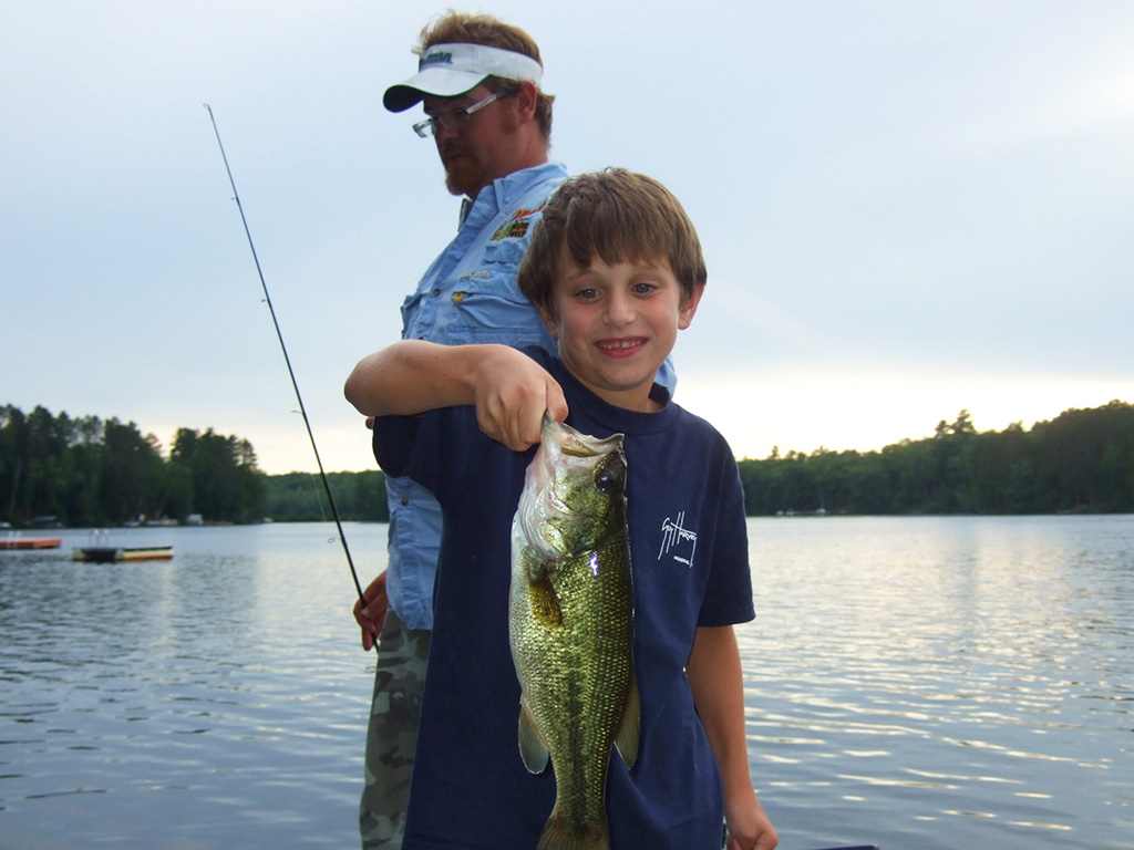 A young angler holds up a Largemougth Bass he recently caught with another angler in the background.