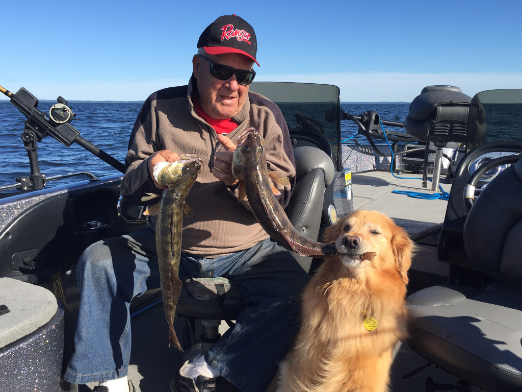 A sitting angler holds up a pair of fish he caught while his dog is biting one of them from behind.