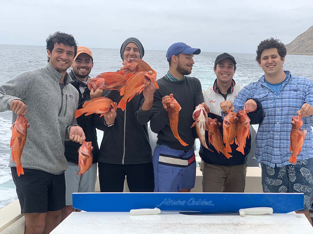 A group of six anglers posing for a photo on a boat, each holding up a pair of Rockfish they caught.