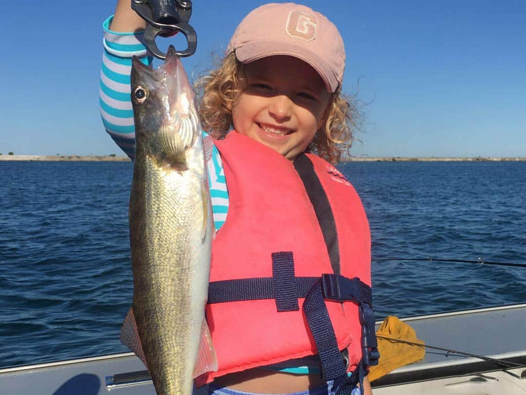 A photo of a happy kid wearing a pink cap and a matching pink life vest while proudly holding a fish in one hand