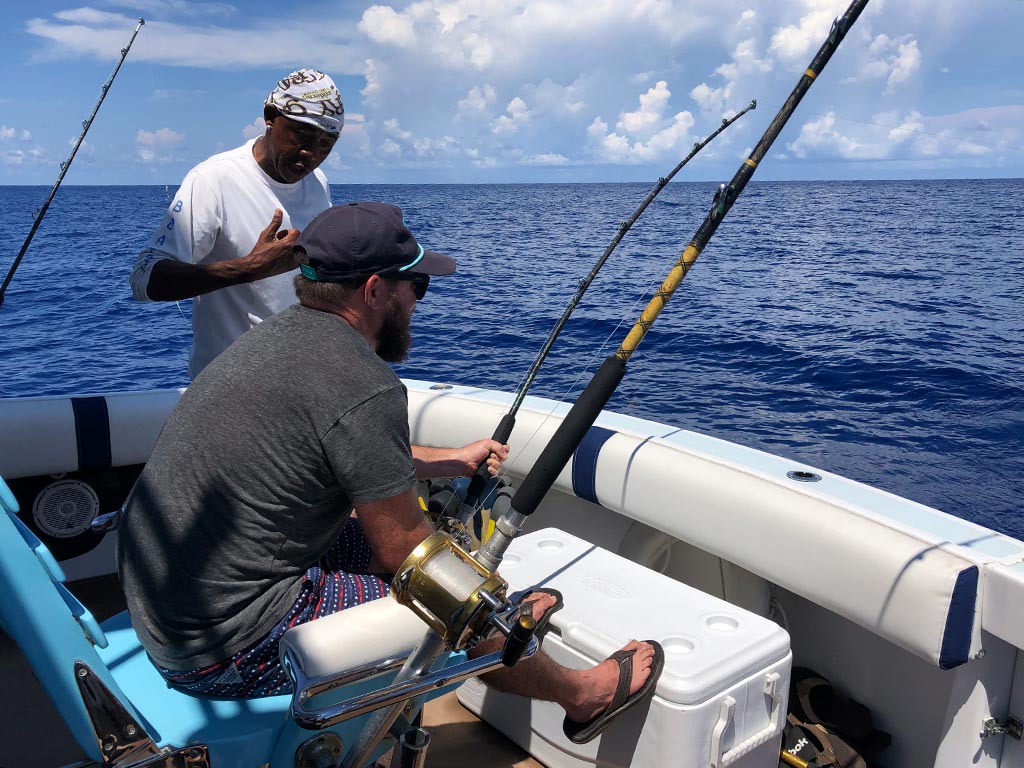 An angler sits on a fighting chair while reeling in a fish, with a crew mate next to him.