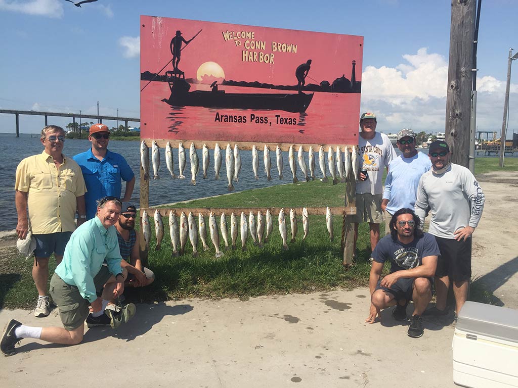 A group of anglers pose around a board stating "Welcome to Conn Brown Harbor" and showing off the numerous Speckled Trout they caught, with the water and a bridge behind them