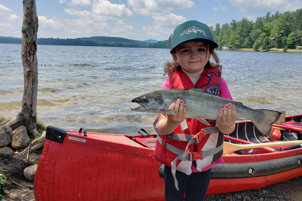 A small girl standing on the shore of a lake, holding an Atlantic Salmon