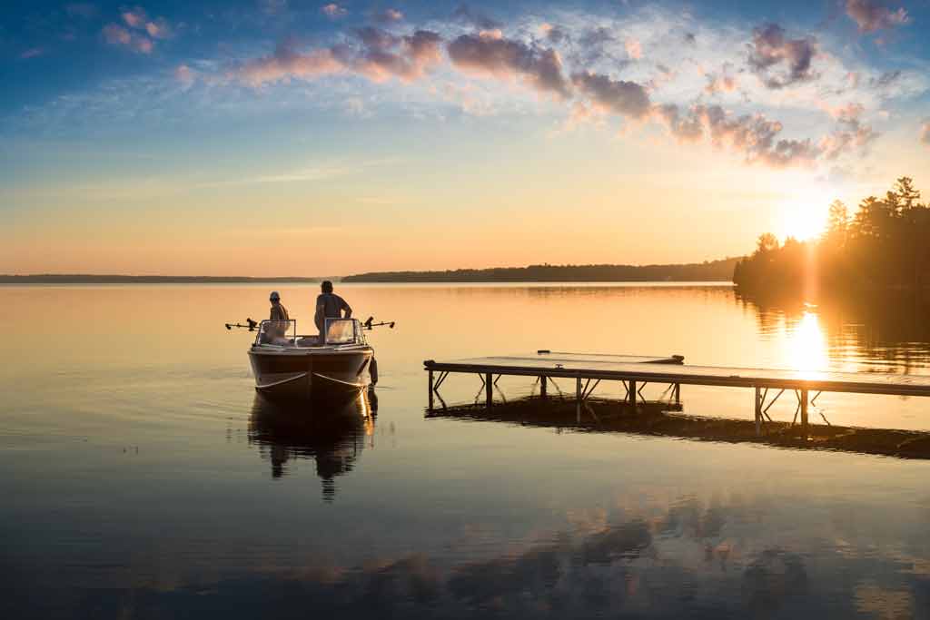 Two people sitting on a fishing boat on a lake, with a dock next to them, at sunset