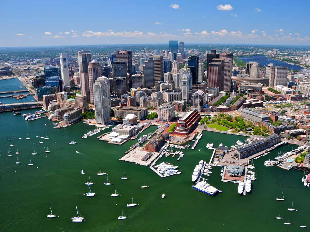 An aerial photo of the city of Boston and numerous boats positioned close to its shores.