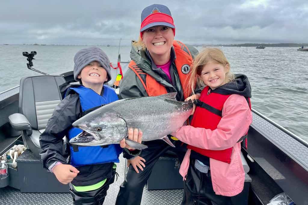 A smiling woman and two kids standing on a boat, holding a Chinook Salmon