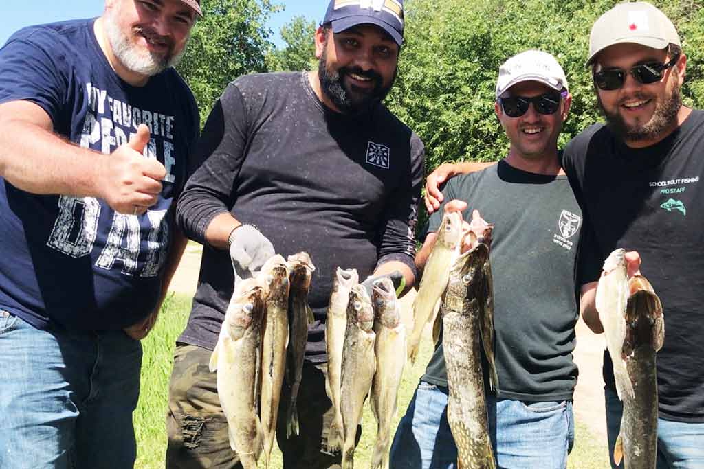 Four men in caps, standing with trees behind them, holding Walleye and Pike they caught