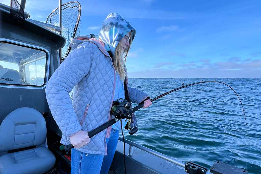 A young woman standing on a fishing boat, holding a fishing rod that's bent because there's a fish on the line