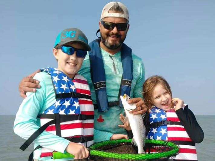 A father and two kids wearing matching American flag life vests, while also holding a Speckled Trout just caught in Texas