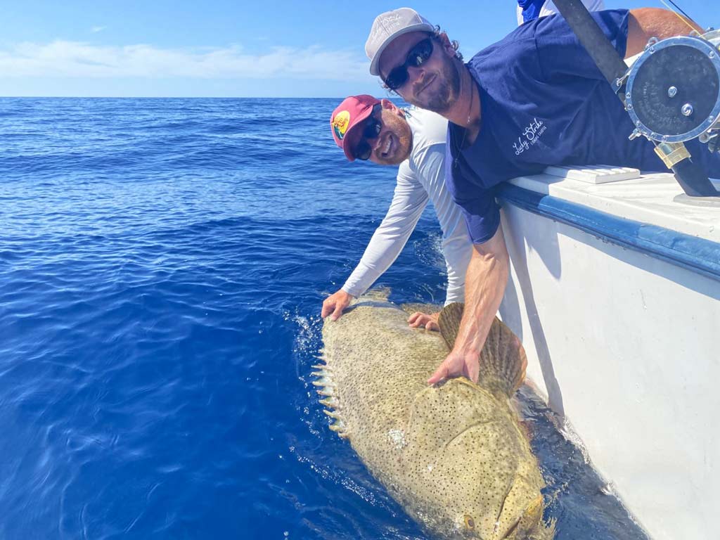 Two anglers leaning over the side of a boat, holding a Goliath Grouper they caught while deep sea fishing in Marco Island.