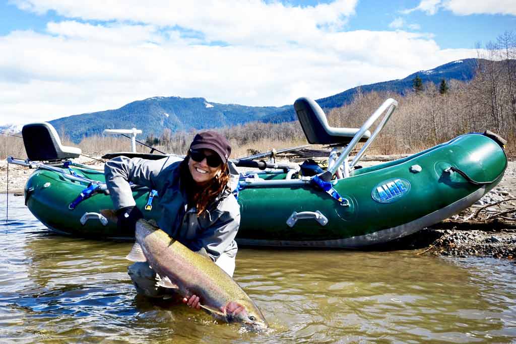 A smiling woman in a cap and sunglasses standing in water, with a boat behind her, holding a big Rainbow Trout