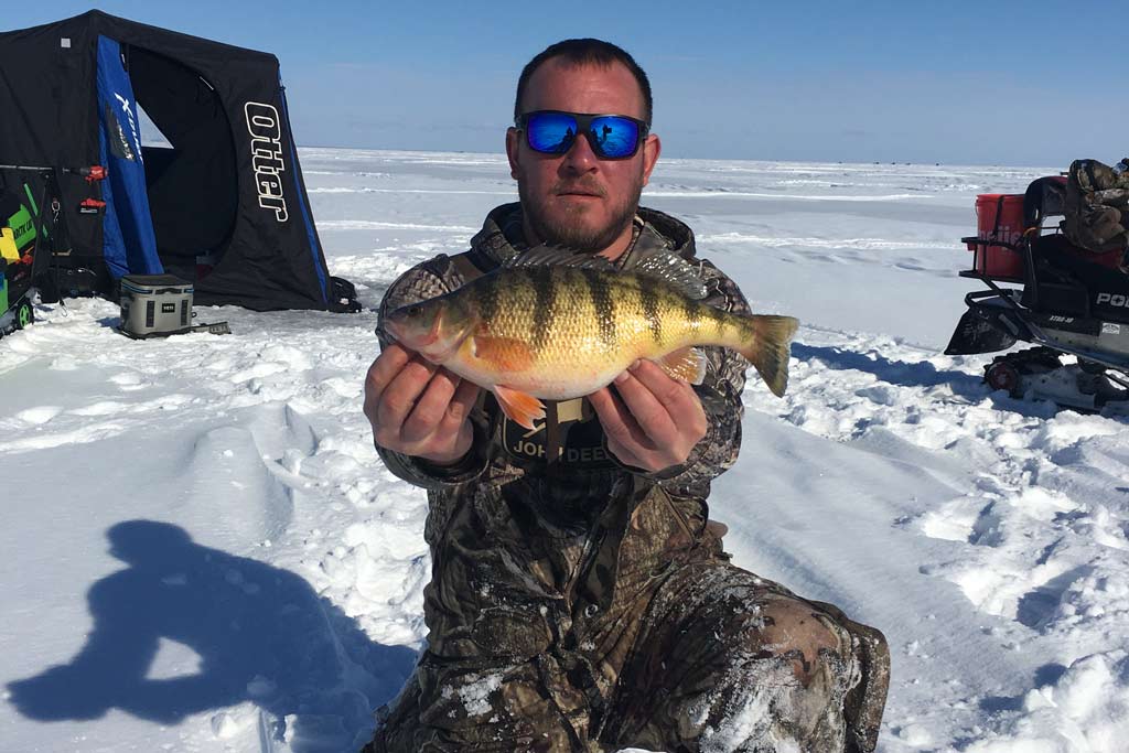 A man in sunglasses holding a Yellow Perch with the frozen Saginaw Bay overdue him, a snowmobile and an ice fishing tent