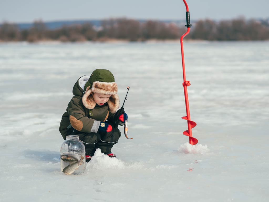 A photo of a kid holding a small rod and bait and squatting on a frozen lake next to a red ice auger