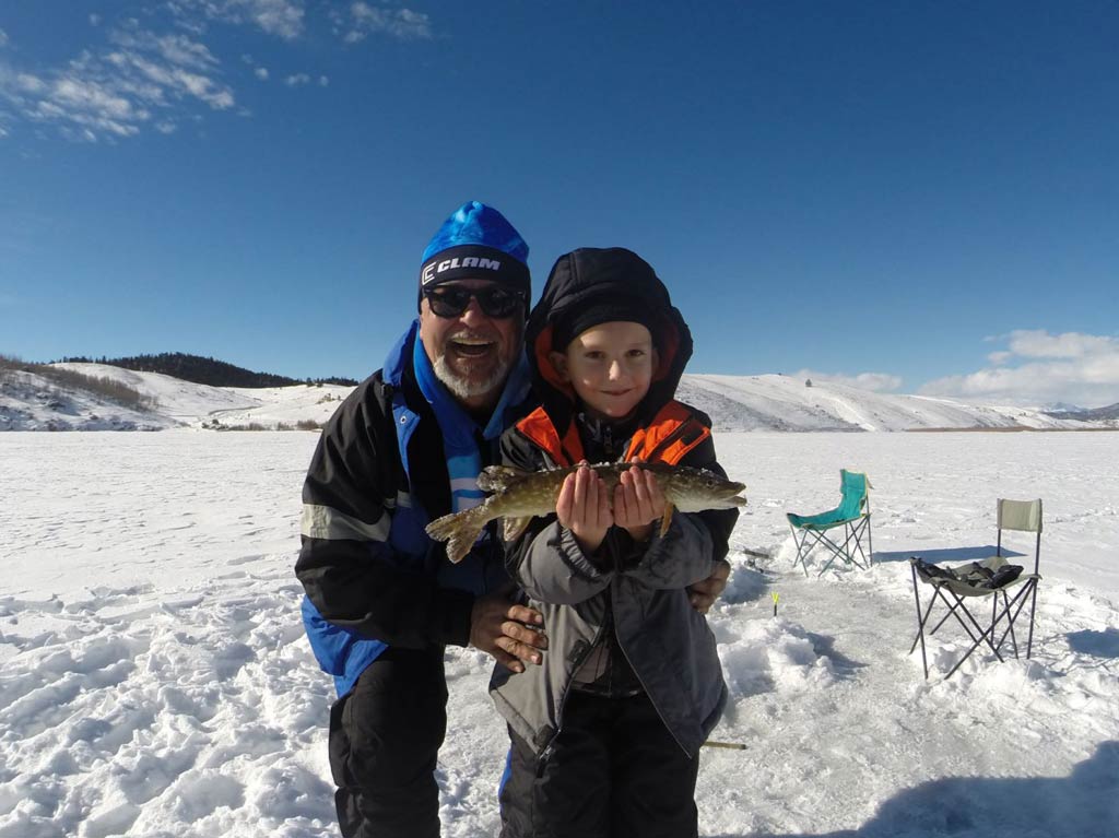 A photo of a dad and his kid on a frozen lake with a small fish caught while ice fishing posing against the chairs and snow-covered hills in the background