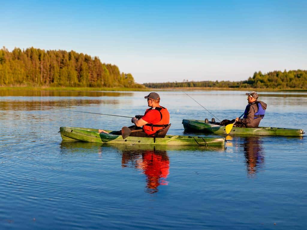 A side view of two anglers fishing from their kayaks on calm lake waters against fall scenery in the background 