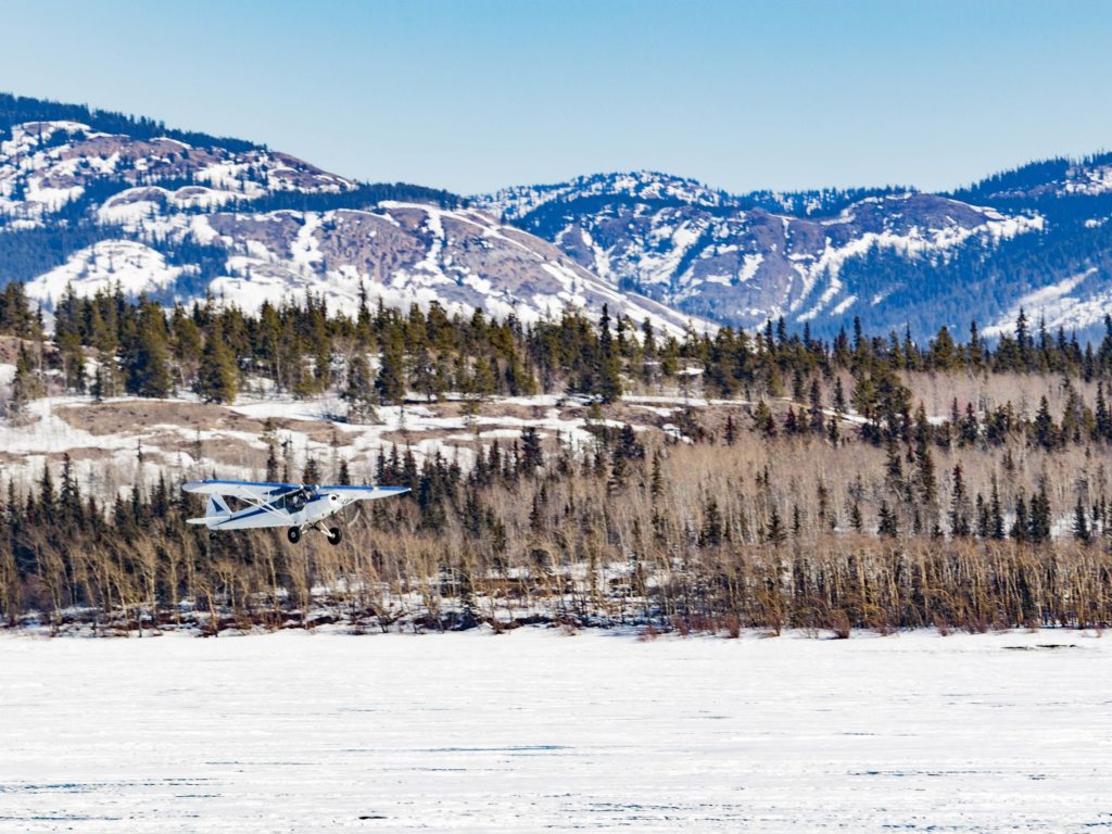 A small aircraft takes off from frozen Lake Laberge in Yukon Territory winter wilderness landscape of boreal forest taiga hills, Yukon, Canada 