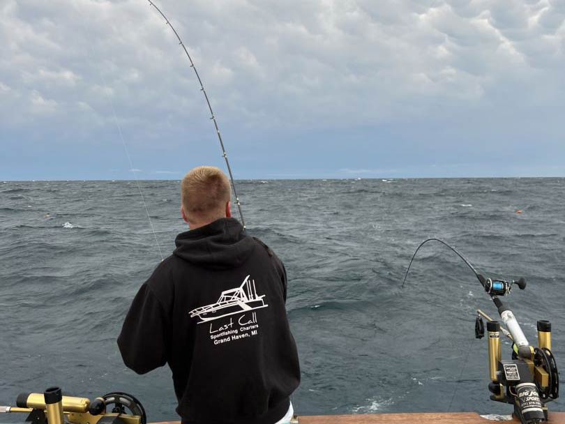 A rear-view picture of the back of a male angler holding a fishing rod while fishing and standing on a charter fishing boat in Grand Haven, MI