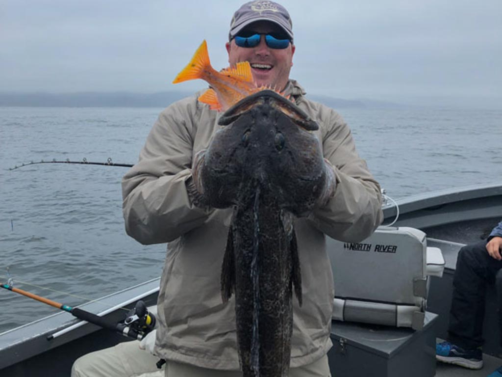 A smiling angler holds up a Lingcod with both of his hands on a cold day.