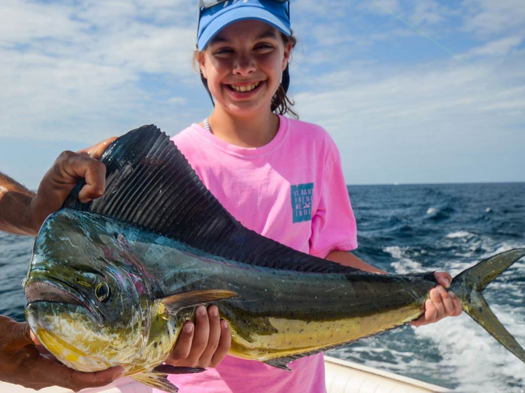 A nice photo of a girl wearing a pink T-shirt and a blue cap and holding big Mahi Mahi with a captain’s helping hand