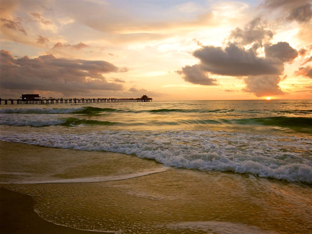 A scenic sunset photo of a beach and the ocean on Marco Island, with a pier on the left-hand side of the image.