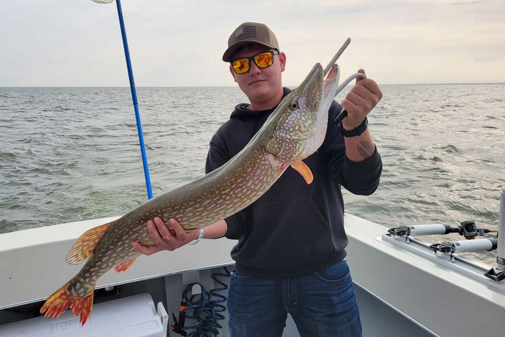 A man in a cap and sunglasses standing on a fishing boat, holding a Northern Pike with water and grey skies in the background