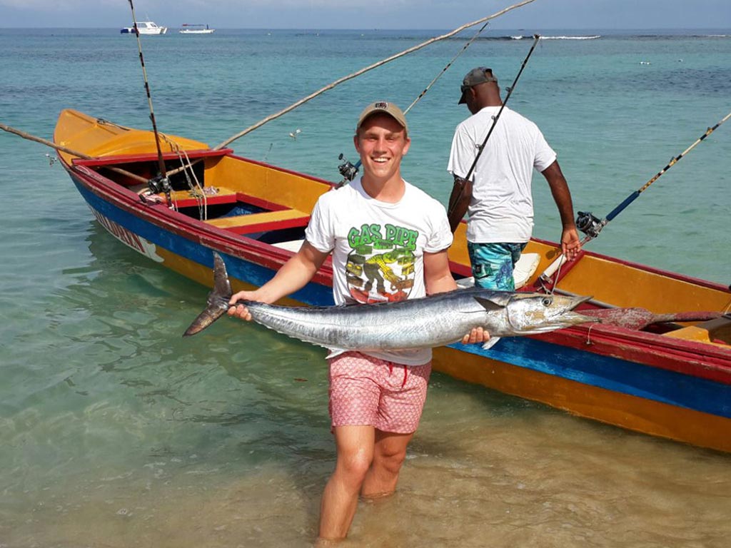 An angler in the surf holds a Wahoo he just caught in Ocho Rios, with a small, wooden fishing boat behind him.