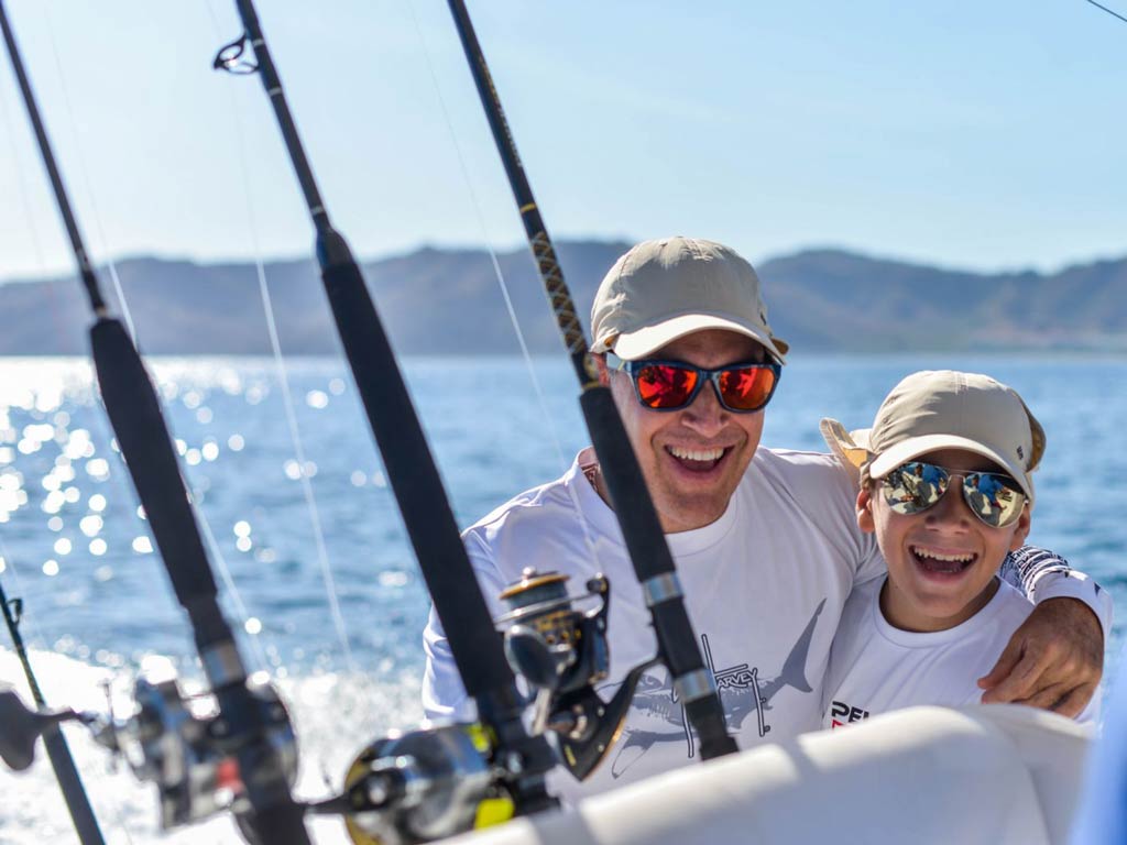 A photo of a father and son hugging and smiling while standing behind rods on a Coco fishing charter boat