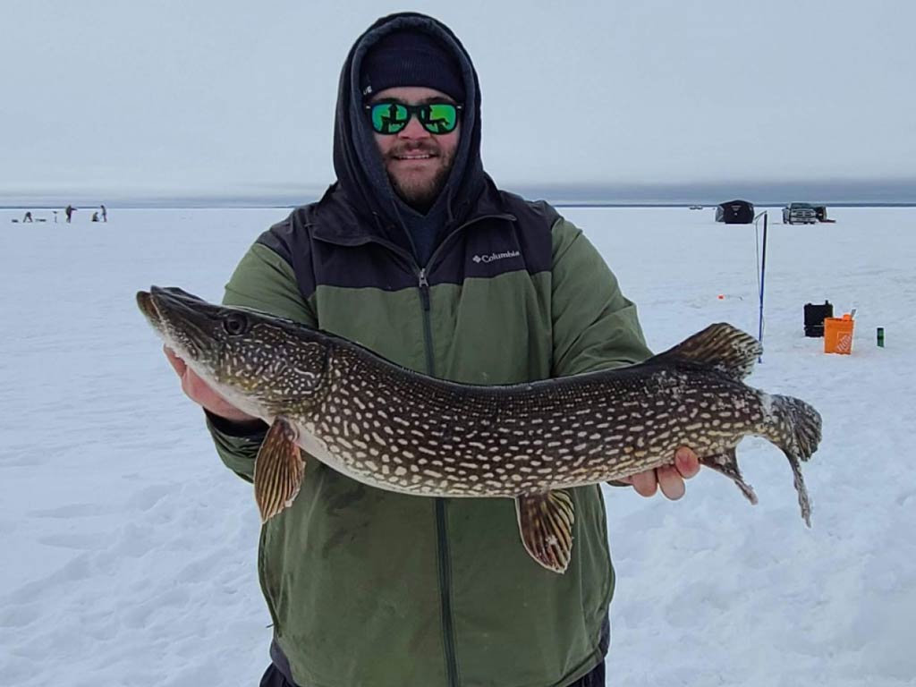 A photo of an angler standing on a frozen lake and holding big Pike