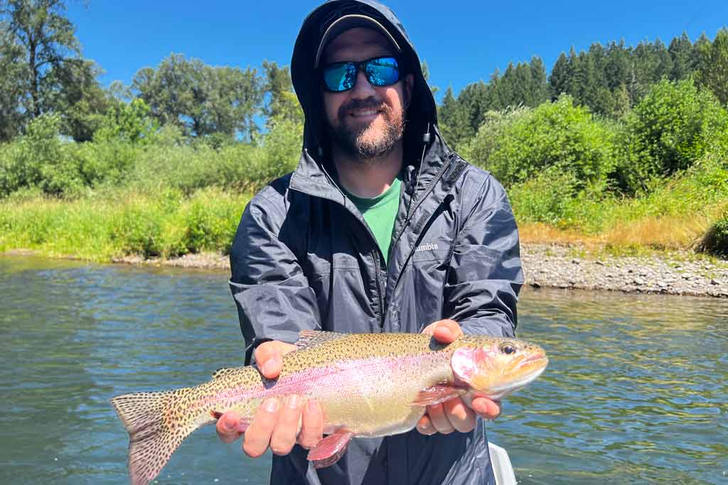 A man in sunglasses, with a hood on, holding a Rainbow Trout, with greenery and a river behind him