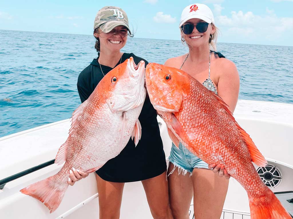 Two women standing on the deck of a charter boat, holding two big Red Snappers they caught deep sea fishing in Marco Island.
