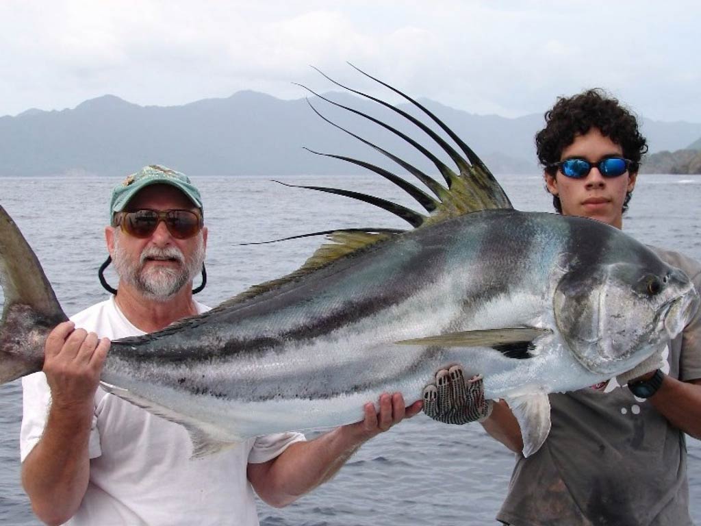 A beautiful side view of Roosterfish and two anglers holding it while standing near the Coco’s shoreline