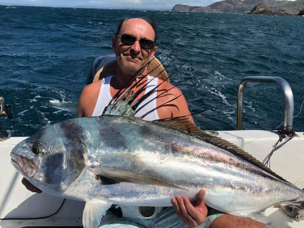 A photo of an older angler sitting on a Coco fishing charter boat and holding big Roosterfish with both hands