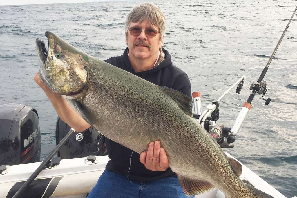 A middle-aged man sitting on a fishing boat, holding a big Chinook Salmon