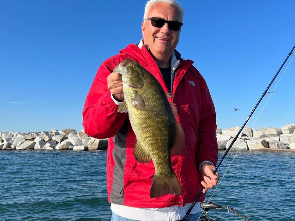 A photo of an angler standing on the shore and holding his catch in one hand and fishing rod in the other, while posing against the jetties in the background