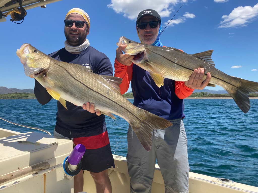 Two Costa Rican anglers standing on a boat, each holding a big Snook they caught.