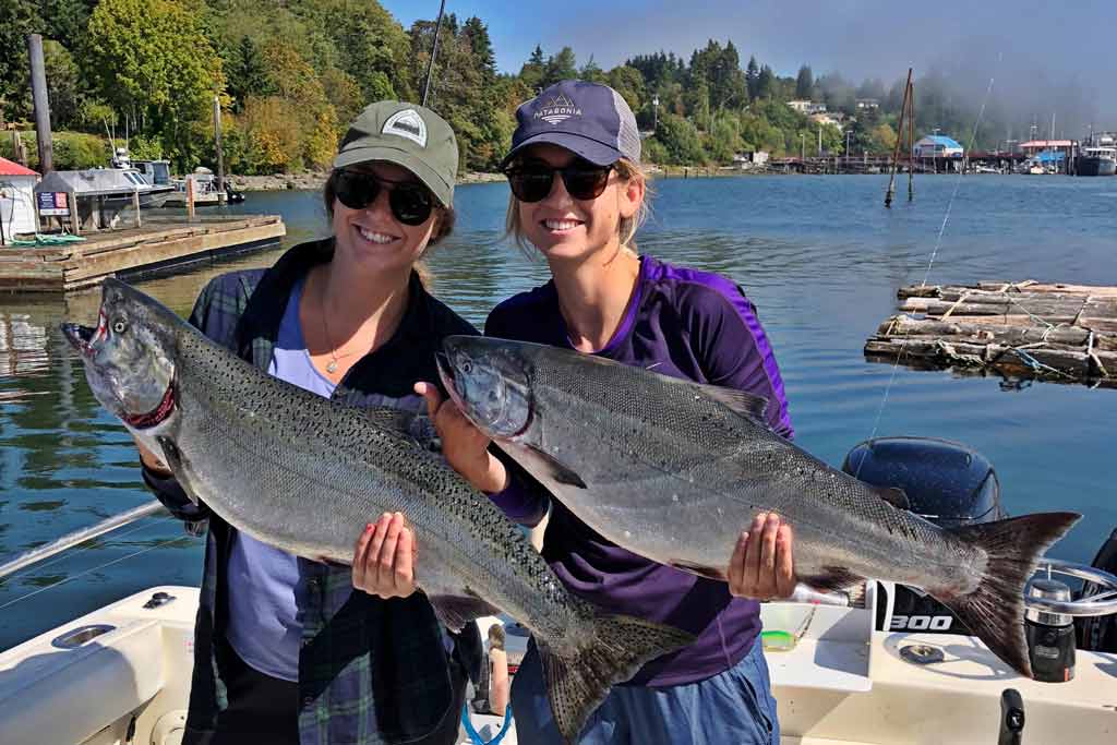 Two smiling women in caps and sunglasses holding two big Chinook Salmon while standing on a fishing boat