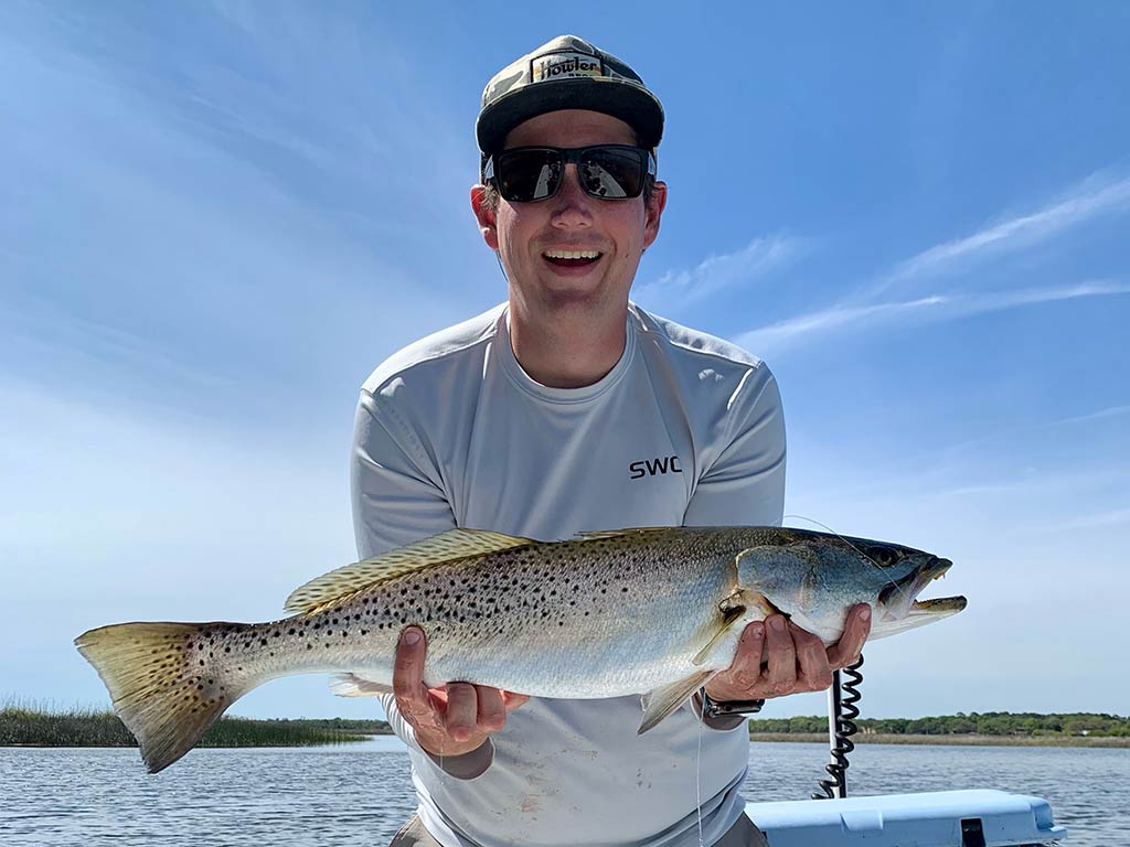 A smiling angler wearing a cap and shades holding a Speckled Trout on a boat