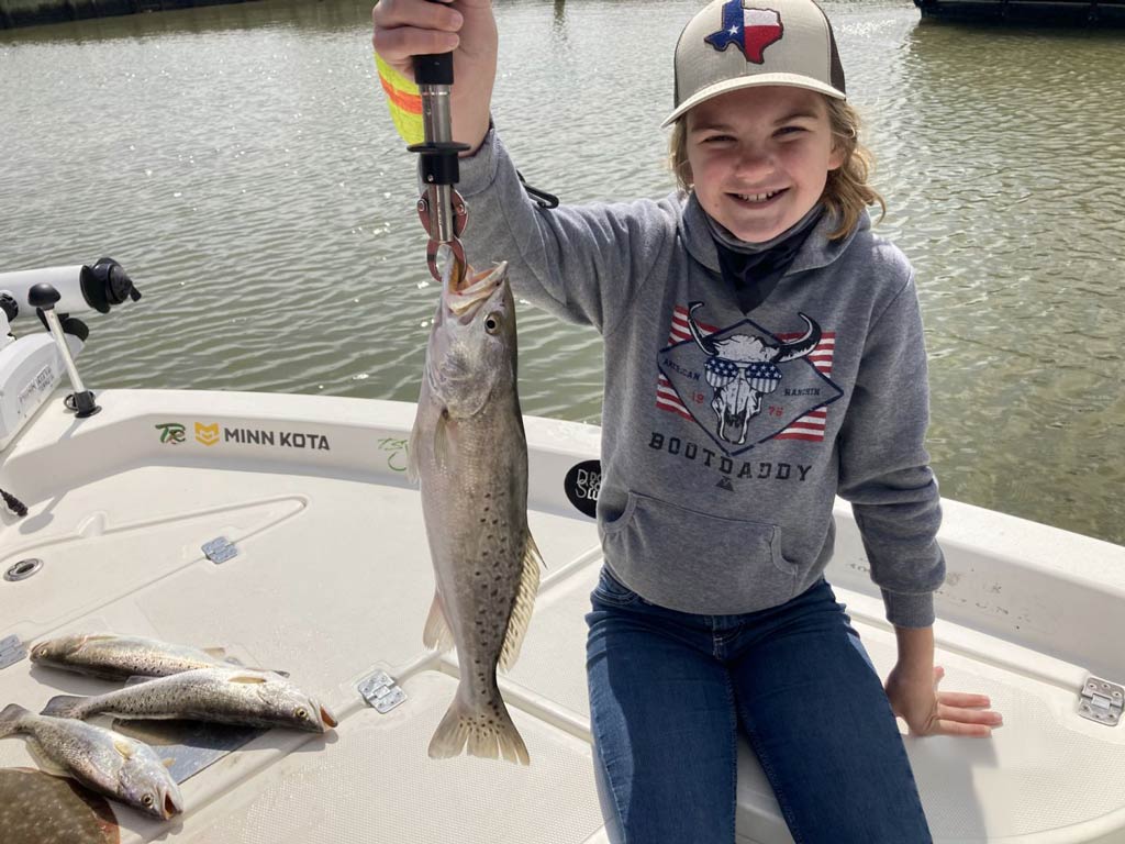 A kid smiling and sitting on a charter boat next to several Speckled Trout and Flounder catches and holding one Speck in his right hand