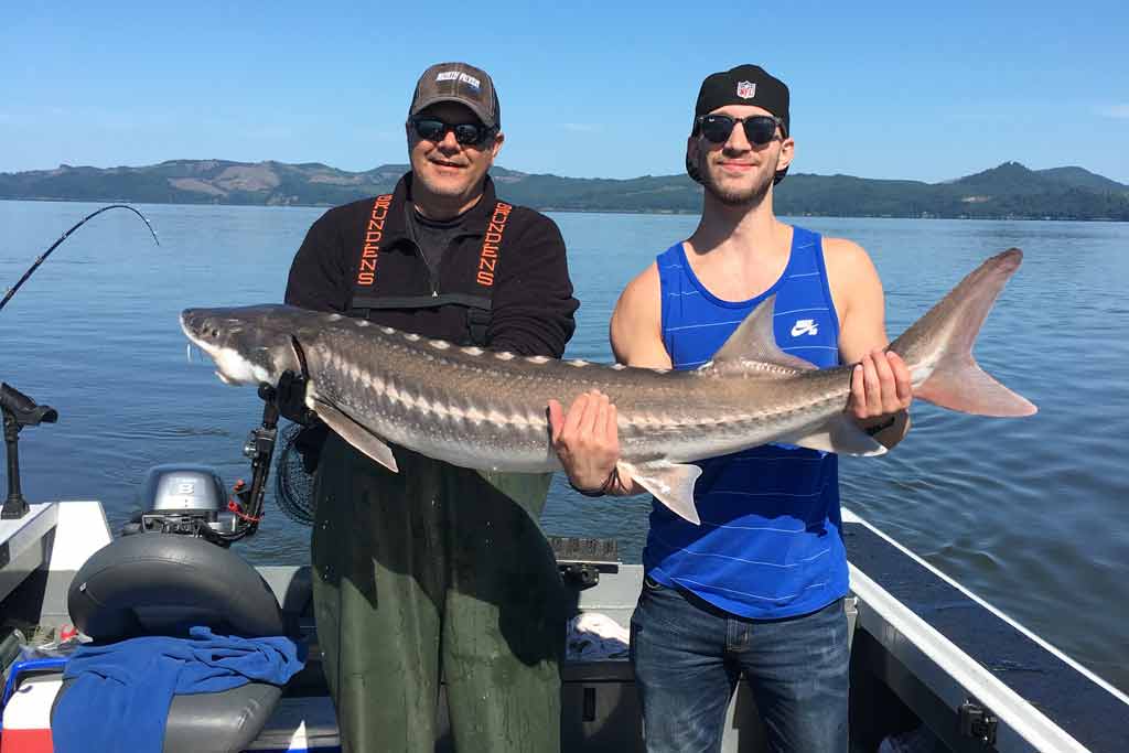 Two men standing on a boat, holding a Sturgeon they just caught, with water and blue skies in the background