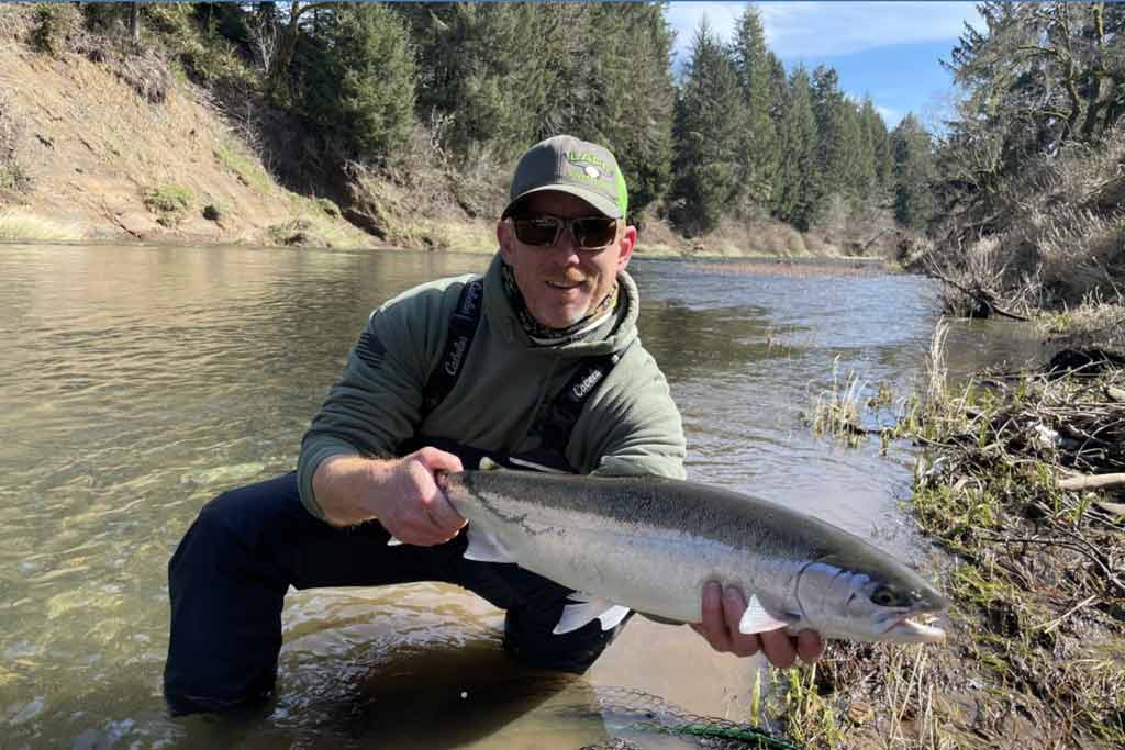 A man in a cap and sunglasses standing in shallow water of a river, holding a Steelhead