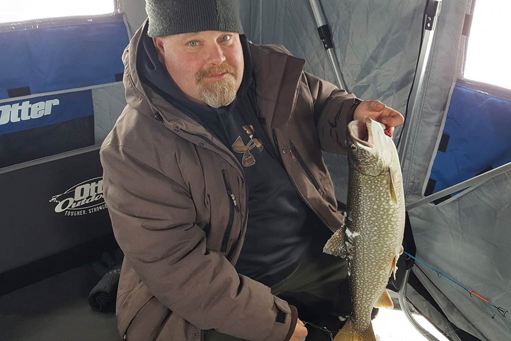 A man in warm winter clothing sitting in an ice fishing tent, holding a Lake Trout