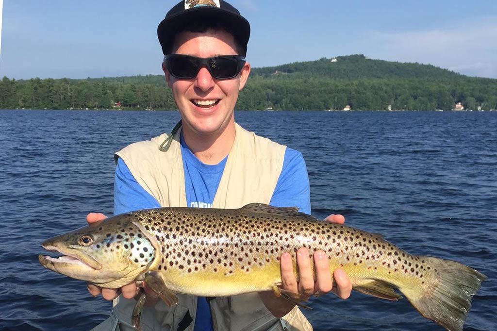 A young angler in sunglasses and a cap, holding a Brook Trout with water in the background