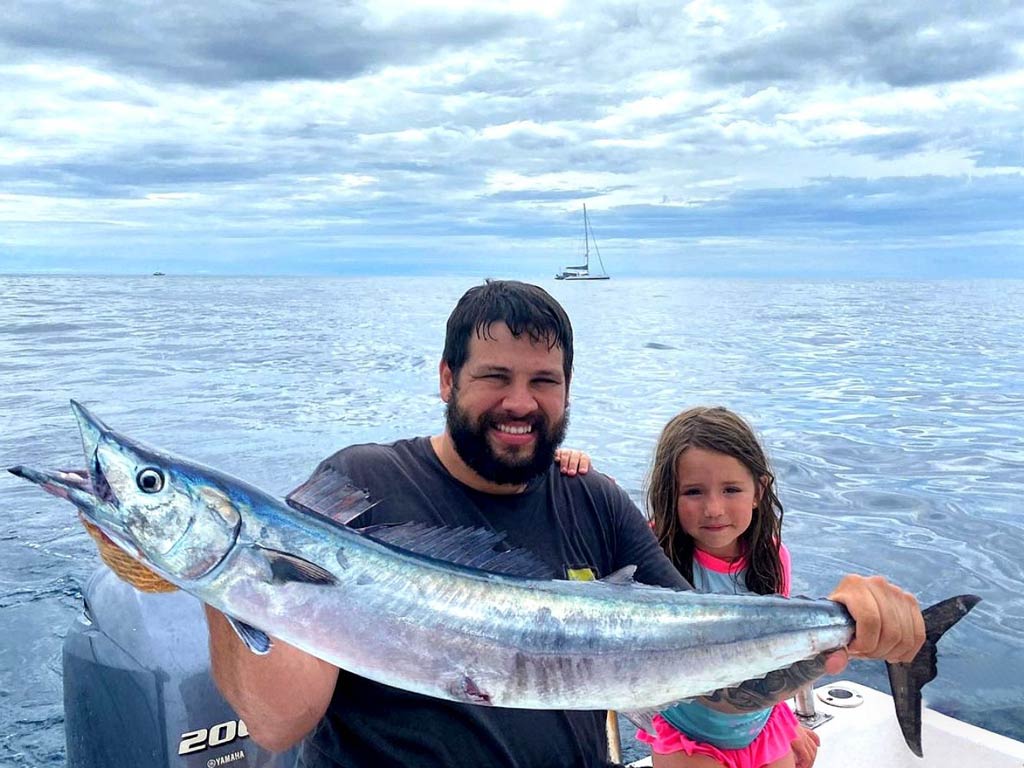 A photo of a father and his daughter sitting on a Coco fishing charter boat and holding Wahoo fish caught while angling in Playas del Coco during a cloudy day