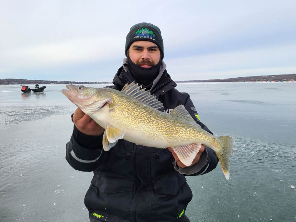 A photo of an angler holding Walleye with both his hands while standing on a frozen lake