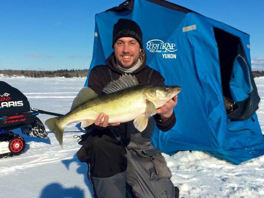 A photo of an angler holding Walleye with both hands while posing on the frozen lake against ice fishing tent and equipment in the background