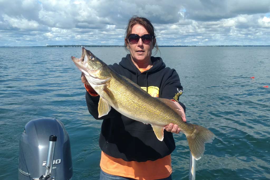 A woman in sunglasses holding a big Walleye while standing on a boat, with water and cloudy skies in the background